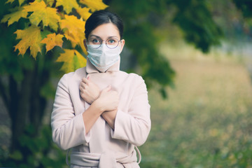 A young woman in a face mask and glasses for vision, wrapped herself in a coat and looks in front of her with eyes wide open. Toned