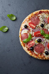 Pizza with salami, mozzarella cheese, cherry tomatoes, black olives, red onion and oregano. Home made food. Concept for a tasty and hearty meal. Black stone background. Top view. Copy space