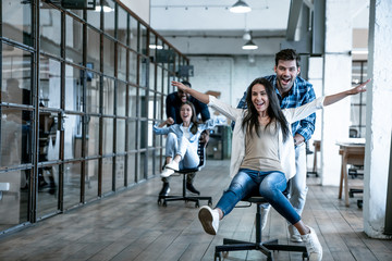 Work hard play hard. Four young cheerful business people in smart casual wear having fun while racing on office chairs and smiling - obrazy, fototapety, plakaty
