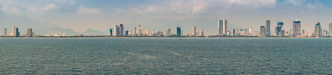 Naklejka premium Nha Trang, Vietnam - March 11, 2019: Panorama shot over Nha Trang bay to skyline of the city under blue cloudscape. Line of tall and shorter buildings. Yellow line of beach.