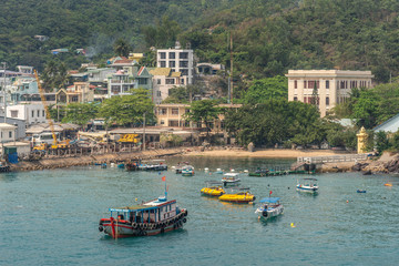 Fototapeta premium Nha Trang, Vietnam - March 11, 2019: Ferry landing pier with colorful small boats in marina adjacent to commercial port. Hotels and housing against green hill flank. 