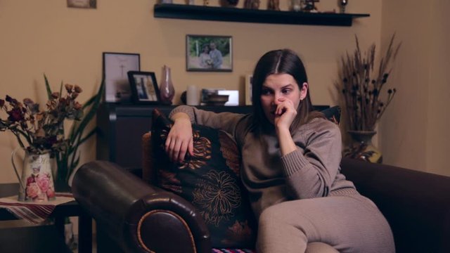 A young pregnant girl of European appearance in a brown suit sits at home in an armchair and picks a finger in her nose