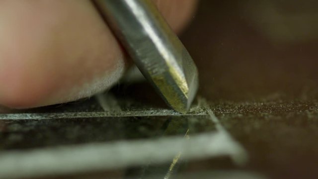 Macro view of process of manual engraving letters on surface of dark granite block as part of headstone, some worker's hand is using metallic tool for this purpose.