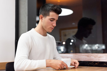 Young man at coffee shop in early morning with a white coffee mug at table .