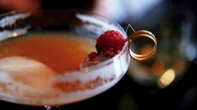close hand held shot with slow focus shift of an orange cocktail with raspberry on a golden stirer in a coupe glass in a dark moody night surrounding bar atmosphere. Packshot with copy space.