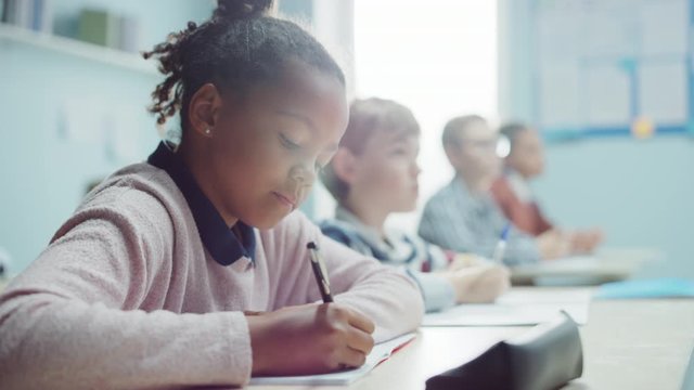 In Elementary School Class: Portrait of Brilliant Girl with Braces Writes in Exercise Notebook, Smiles. Junior Classroom with Diverse Group of Bright Children Working Diligently and Learning New Stuff