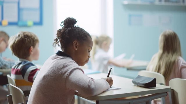 In Elementary School Class: Portrait of a Brilliant Black Girl with Braces Writes in Exercise Notebook, Smiles. Junior Classroom with Diverse Group of Bright Children Working and Learning New Stuff