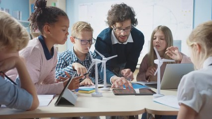 Elementary School Classroom: Enthusiastic Teacher Holding Tablet Computer Explains to a Brilliant Young Children How Wind Turbines Work. Kids Learning about Eco-Friendly Forms of Renewable Energy - Powered by Adobe
