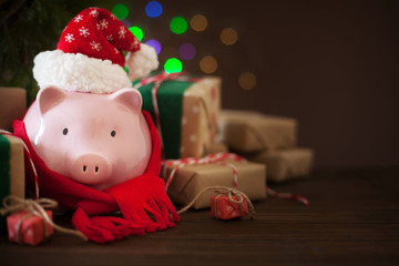 Piggy bank in santa claus christmas hat. Gift boxes, spruce branches, and a garland.