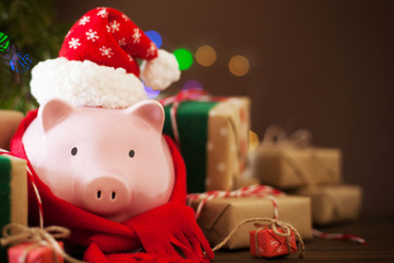 Piggy bank in santa claus christmas hat. Gift boxes, spruce branches, and a garland.
