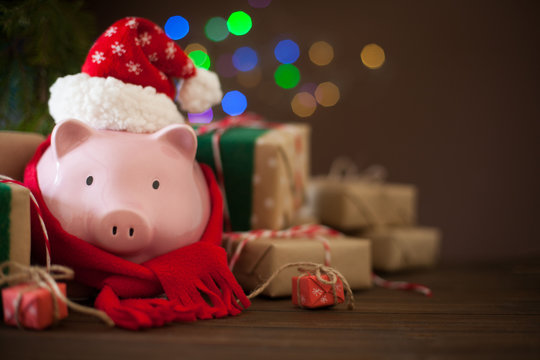 Piggy bank in santa claus christmas hat. Gift boxes, spruce branches, and a garland.