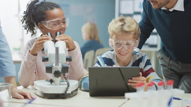 Elementary School Science Classroom: Cute Little Girl Looks Under Microscope, Boy Uses Digital Tablet Computer to Check Information on the Internet, while Enthusiastic Teacher Explains Lesson
