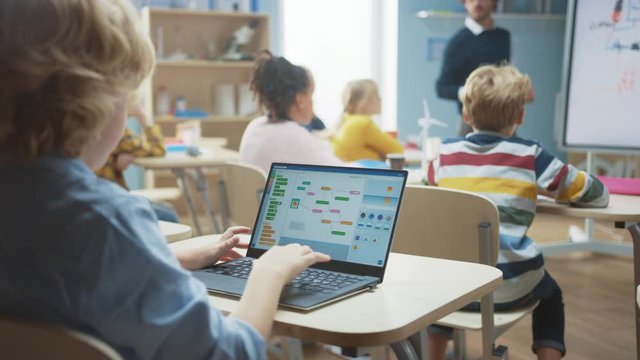 Elementary School Science Class: Over the Shoulder Little Boy Uses Laptop with Screen Showing Programming Software. Physics Teacher Explains Lesson to a Diverse Class full of Smart Kids