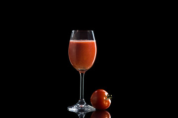 a glass of tomato juice stands on a reflective surface on a dark background and next to it lies a red round tomato.