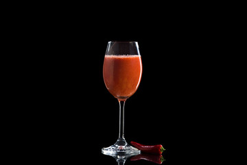 a glass of wine with tomato juice stands on a reflective surface on a dark background next to it lies red chili pepper.