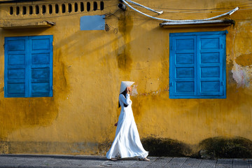 Young Vietnamese Woman in White Tunic  Walking in Front of a Weathered Gold Stucco Wall with Blue Shutters