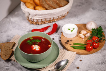 Tasty and hearty dinner. A plate with borsch on the table, next to the board are vegetables, parsley, dill, green onions, garlic, chili peppers, cherry tomatoes, and bread.