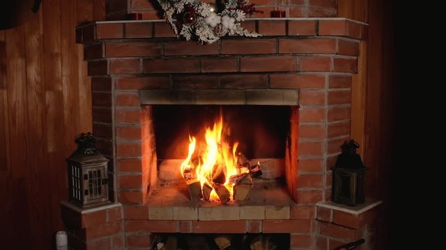 flame of fire burns in brick home Christmas fireplace. festive wreath hangs over fireplace. Wooden candlesticks is decorated with Christmas decor. Rest and relaxation in front of fireplace in evening.
