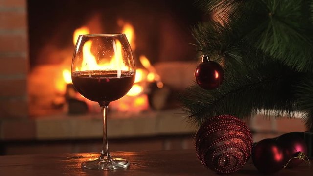 red wine in glass stands on table against background of burning flames of Christmas fireplace. Christmas artificial Christmas tree with beautiful decorations. New year cozy holidays with family