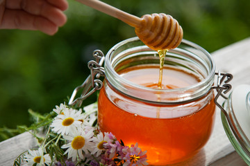 Honey in glass jar with bee flying and flowers on a wooden floor.