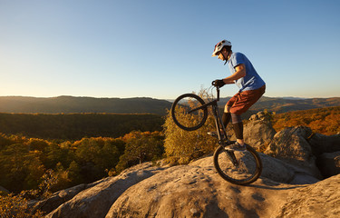 Fototapeta na wymiar Young cyclist balancing on back wheel on trial bicycle. Sportsman rider making acrobatic trick on the edge of big boulder on the top of mountain at sunset. Concept of extreme sport active lifestyle