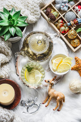 Glass teapot with green tea, flower, candle, knitted plaid and box of christmas decorations on a fluffy carpet on a light background. Christmas background