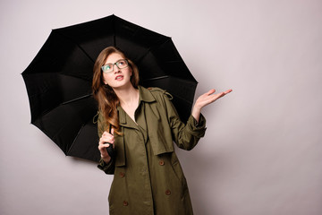 A girl stands on a light background in a green trench coat and glasses under a black umbrella and looks up, checking the rainfall