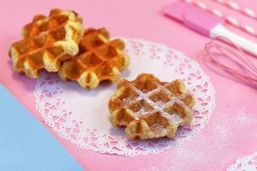 Fresh small round waffles with sugar powder on paper lace doily and pink ground with baking utensils in background