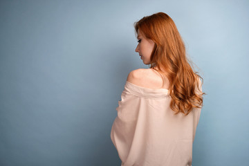 Fototapeta premium The red-haired girl stands with her back in a light robe with a bare shoulder, her head bowed against a blue background.