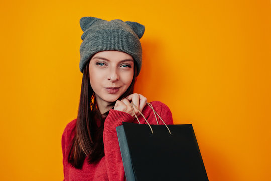 Woman in gray hat on the orange background. Young woman in sweater and hat holds black bag. Black friday image. Woman shopping.