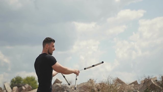Handsome, young man with a beard in a black T-shirt demonstrates mastery with nunchaku outdoors on a stone background.