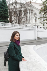 Beautiful young stylish brunette woman with short haircut, wearing green coat, black backpack, purple scarf walking through the city streets. Trendy casual outfit. Everyday look. Street fashion.