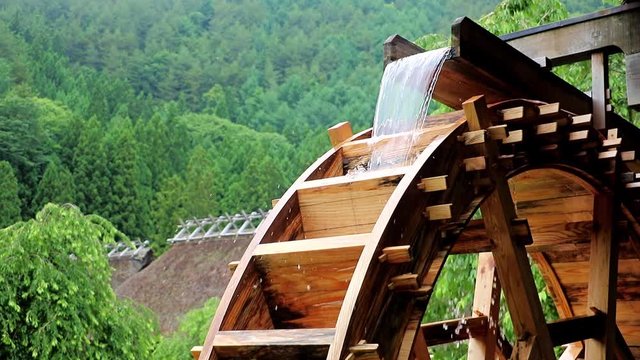 The mill wheel rotates under a stream of water at village with traditional thatched roofed houses, Japan.