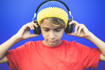 Portrait of a beautiful boy listening music with headphones isolated on a blue background. Colorful image of a young male wearing a wool cup, have fun with earphones. Youth, fun, isolation concept.