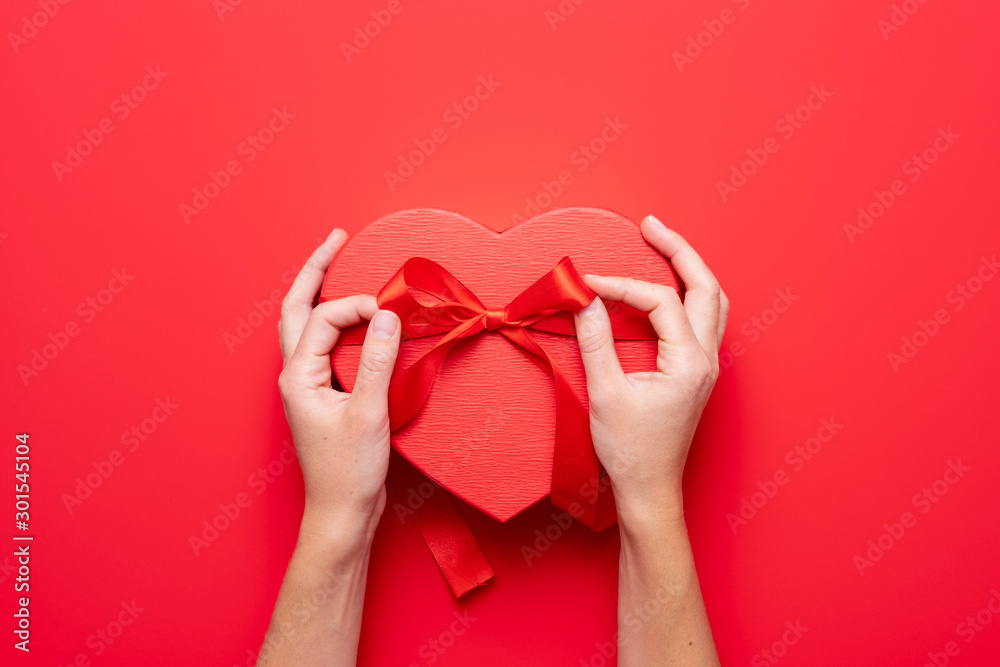 Wall mural Close up on female hands holding a gift in a pink heart presents for valentine day, birthday, mother's day. Flat lay. Symbol of love. Valentines day background with a gift boxes on concrete board.