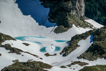 A view on a ice-frozen alpine lake. The ice shines in many different shades of blue. Lake is surrounded by tall mountains. There is no snow on the peaks. Spring slowly coming to high altitudes