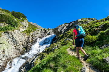 Naklejka na ściany i meble A young woman with a big backpack hikes up on a steep pathway, along a mountain stream. the water cascades on the rocks, rushing into the valley. Girl is enjoying the spring in the mountains