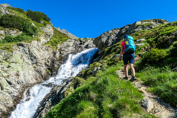Naklejka premium A young woman with a big backpack hikes up on a steep pathway, along a mountain stream. the water cascades on the rocks, rushing into the valley. Girl is enjoying the spring in the mountains