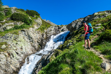Fototapeta premium A young woman with a big backpack hikes up on a steep pathway, along a mountain stream. the water cascades on the rocks, rushing into the valley. Girl is enjoying the spring in the mountains
