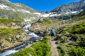 Fototapeta na wymiar A young woman with a big backpack hikes up on a steep pathway, along a mountain stream. the water cascades on the rocks, rushing into the valley. Girl is enjoying the spring in the mountains