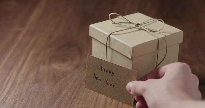 Slow motion man hand put happy new year card next to brown paper gift box on walnut table