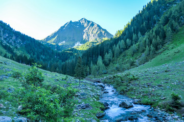 Fototapeta premium A view on Schladming Alps, partially still covered with snow. A small stream flowing between the stones. Spring slowly reaching the tallest parts of the mountains. First sunbeams reaching the peak.