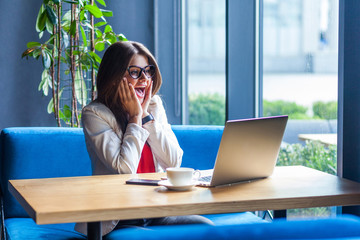 Portrait of surprised beautiful stylish brunette young woman in glasses sitting, reading amazing news on her laptop screen and looking with amazed face. indoor studio shot, cafe, office background.