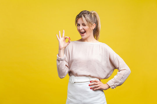 Portrait of satisfied beautiful young woman with blonde hair in casual beige blouse standing, looking at camera showing Ok sign gesture and winking, indoor studio shot isolated on yellow background