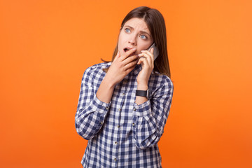 Portrait of worried young woman with brown hair wearing checkered shirt standing talking on smartphone, looking up and having pleasant conversation. indoor studio shot isolated on orange background