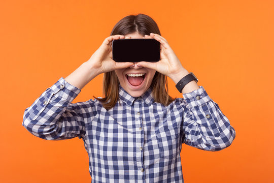 Portrait of happy amazed woman with toothy smile and brown hair wearing checkered shirt standing, hiding her eyes with cellphone, taking pictures. indoor studio shot isolated on orange background