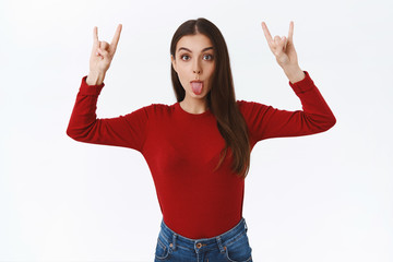 Playful, funny and silly brunette girl in red sweater, raising hands up showing rock-n-roll, heavy metall sign, showing tongue with goody expression, standing white background fool around