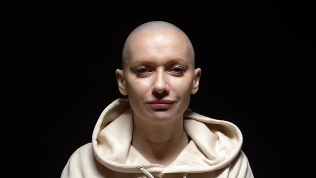 bald woman in a hoodie looks at the camera and smiles sadly. against a dark background.