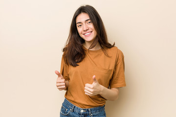 Obraz premium Young brunette woman against a beige background raising both thumbs up, smiling and confident.