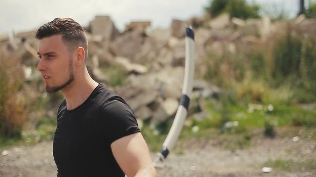 Handsome, young man with a beard in a black T-shirt, looks into the camera and demonstrates mastery with nunchaku outdoors.